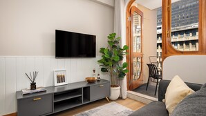 Uma sala de estar moderna com uma TV de tela plana na parede, um rack cinza para entretenimento, uma planta grande em vaso e vista de um prédio por uma porta aberta.