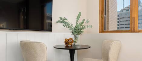 A room with two chairs, a round table, a vase with greenery, and a TV mounted on the wall.