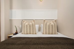 Une chambre d’hôtel comprenant un lit, des oreillers, une table de chevet, un vase avec une plante et un luminaire fixé au mur.