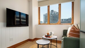 Ein modernes Wohnzimmer mit einem an der Wand befestigten Flachbildfernseher, einem kleinen runden Tisch mit Büchern und einem Dekoelement, einer Couch mit einer Zudecke und einem großen Fenster mit Blick auf Gebäude.