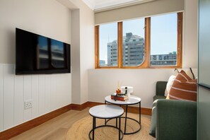 A modern living room with a flat-screen TV mounted on the wall, a small round table with books and a decorative item, a sofa with a throw blanket, and a large window with a view of buildings. - Charlie and the Chairman | Elevated City Stay (Cape Town)