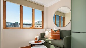 Ein modernes Wohnzimmer mit einem grünen Sofa, einem runden Spiegel, einem kleinen weißen Tisch und Blick auf Gebäude durch große Fenster.