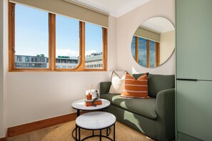 A modern living room with a green sofa, a round mirror, a small white table, and a view of buildings through large windows. - Charlie and the Chairman | Elevated City Stay (Cape Town)