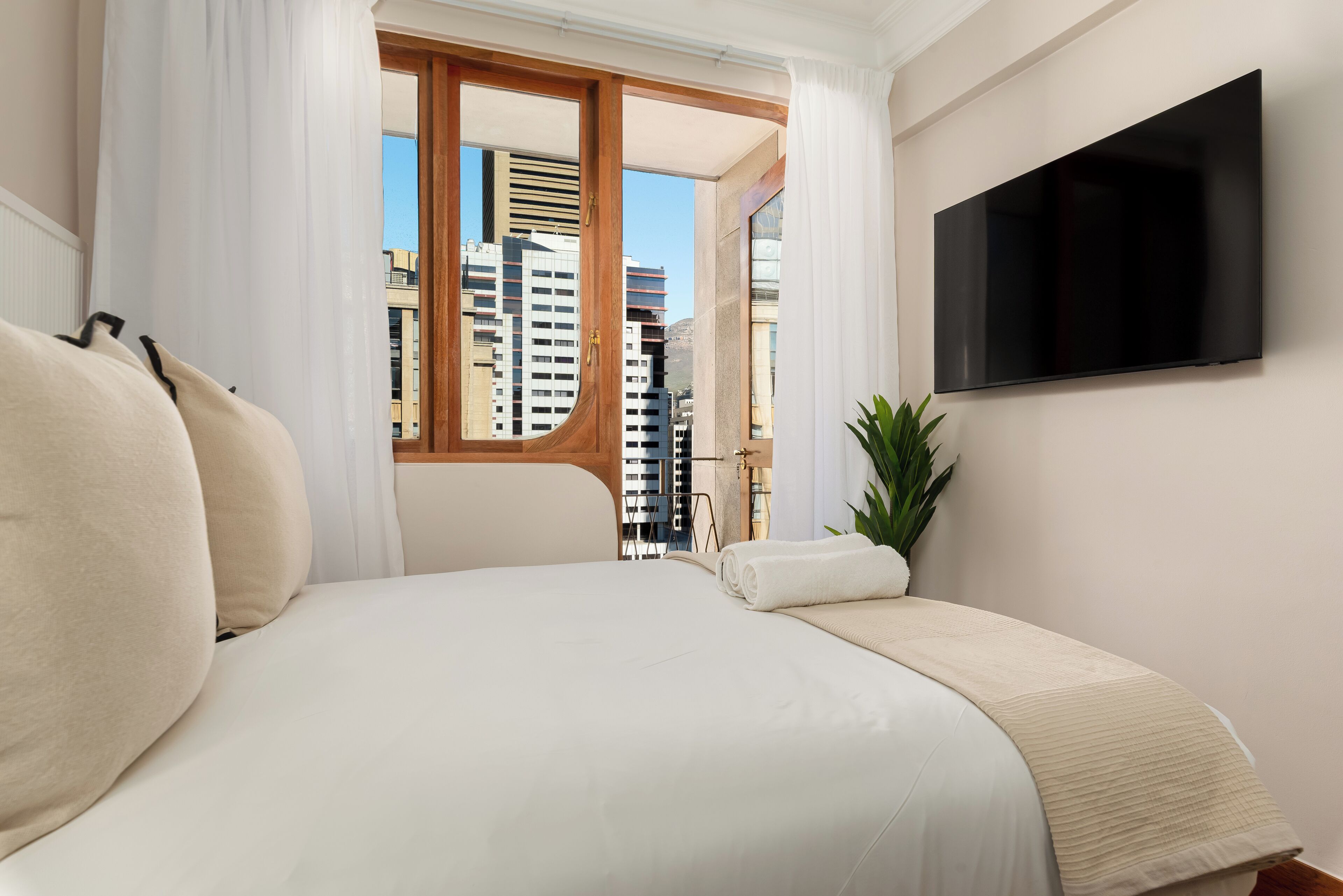 A hotel room with a large bed, a flat-screen TV mounted on the wall, and a view of city buildings through a window.
