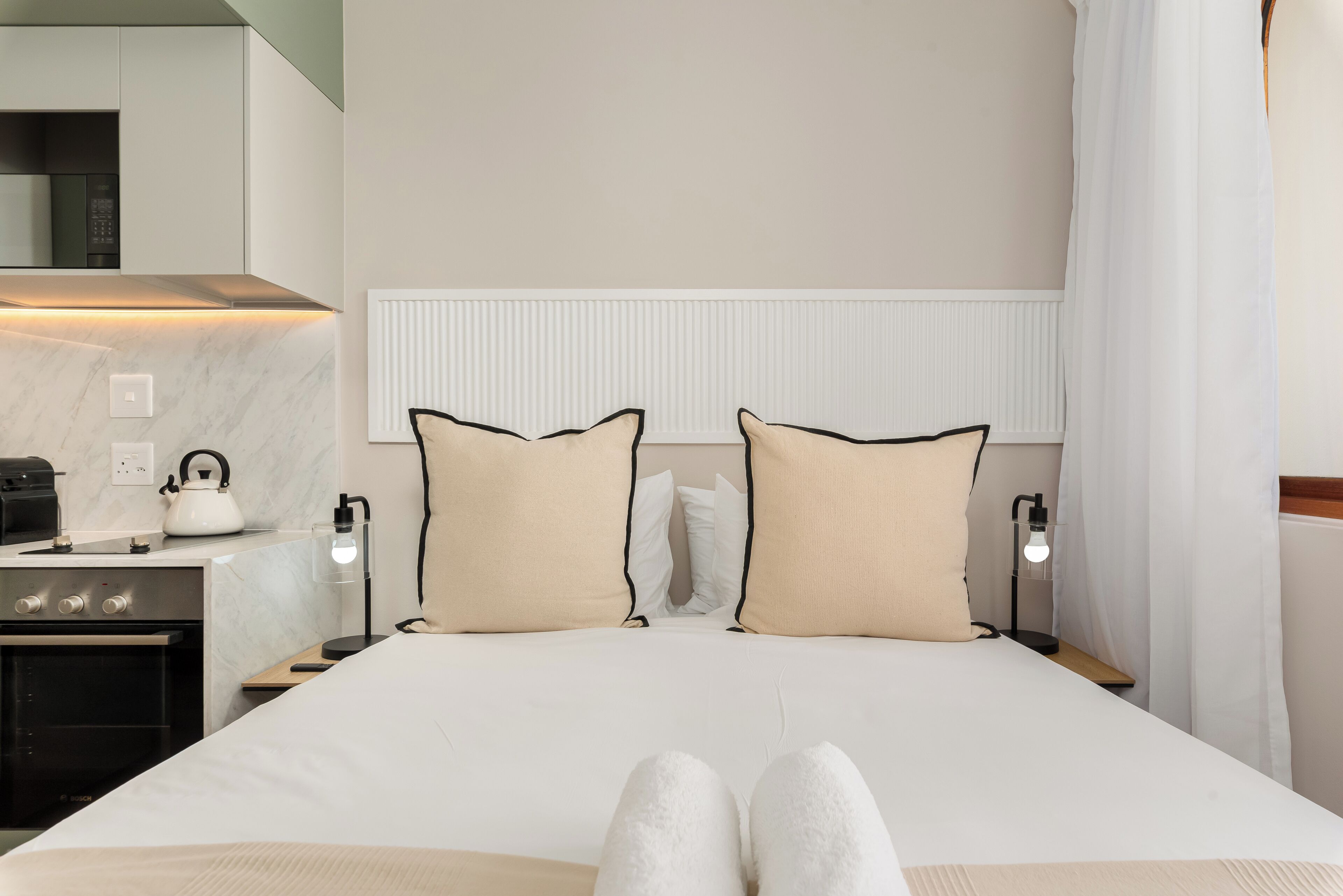 A hotel room with a bed, bedside lamps, a kettle, and a modern kitchen area.