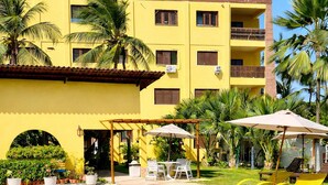 A swimming pool with a curved edge, surrounded by a yellow building with multiple balconies, palm trees, and outdoor seating. - Pousada Sunrise Beach (Caucaia)