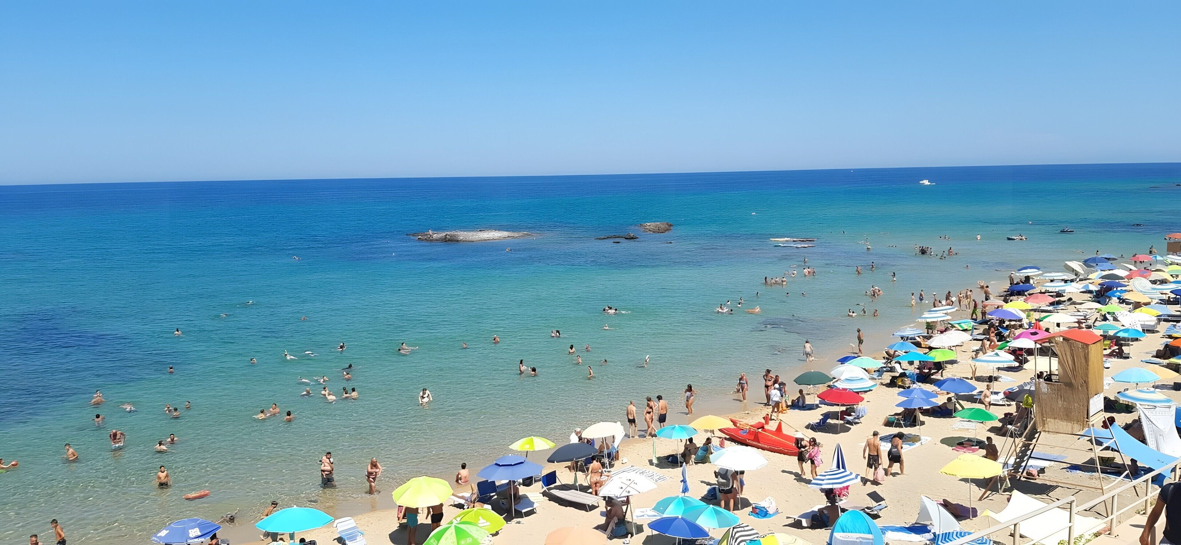 Une plage à l’eau d’un bleu limpide, où l’on peut voir des gens se baigner, et où de nombreux parasols colorés sont installés.