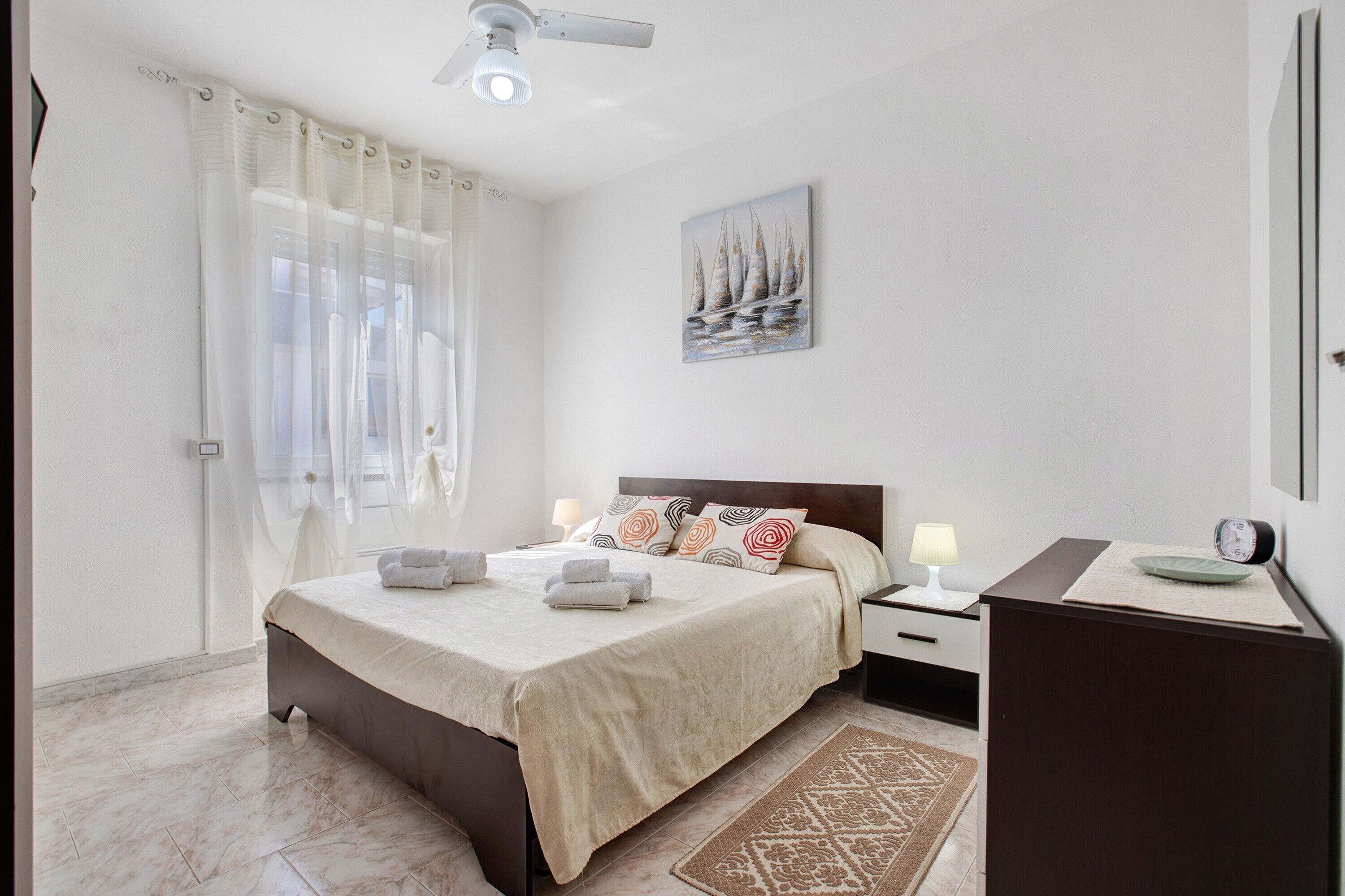 Ein Hotelzimmer mit Bett, Nachttischen, Schreibtisch, Stuhl, Lampe, einer Uhr, einem Bild, Deckenventilator und einem Fenster mit Vorhängen.