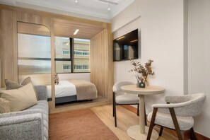 Una habitación de hotel moderna con una cama, dos sillones, una pequeña mesa redonda, un televisor montado en la pared y una ventana con persianas.