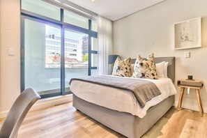 Ein modernes Schlafzimmer mit einem großen Bett, einem Sessel, einem Nachttisch und Blick auf Gebäude durch ein großes Fenster.