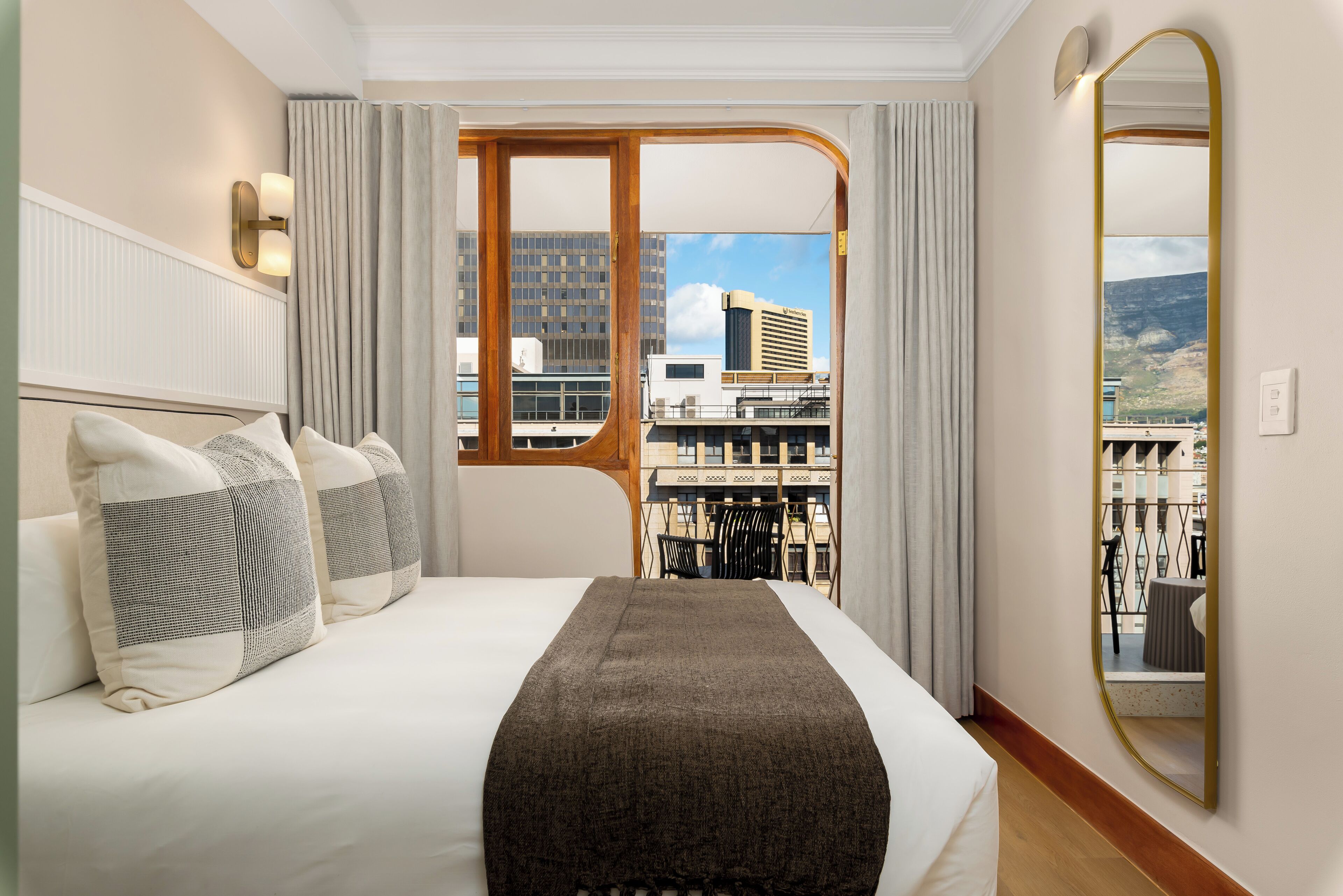 A hotel room with a large bed, a view of the cityscape, a mirror, and a window with curtains.