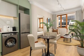 Un salon moderne avec un canapé, une table ronde et une table basse. La cuisine adjacente comprend un lave-linge et un réfrigérateur.