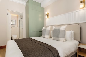 A bedroom with a bed, bedside tables, a nightstand, a wall-mounted light fixture, and a door leading to a bathroom. - Charlie and the Chairman | Refined Living and Views (Cape Town)