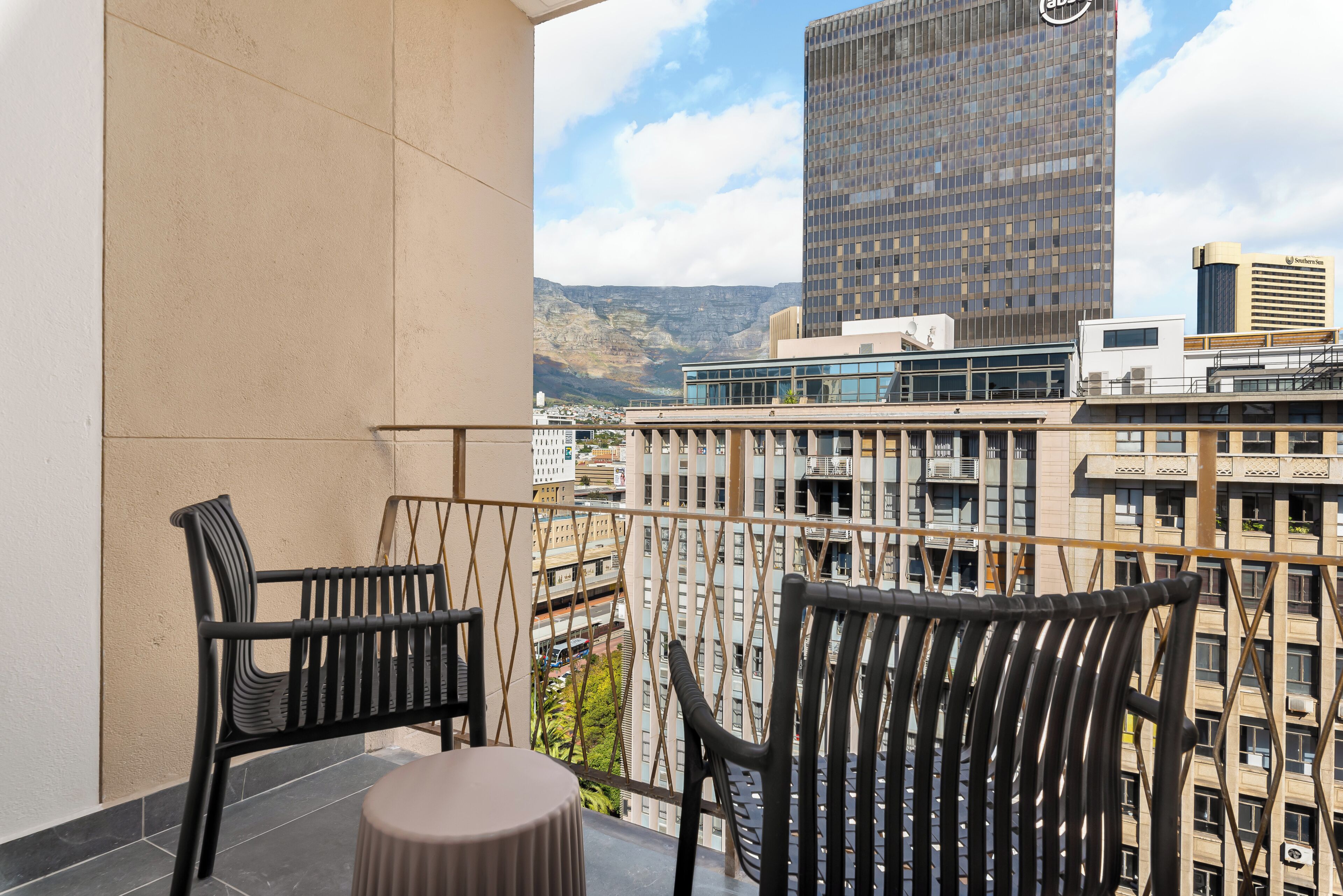 A balcony with two chairs and a small table, overlooking a cityscape with buildings and a mountain in the distance.