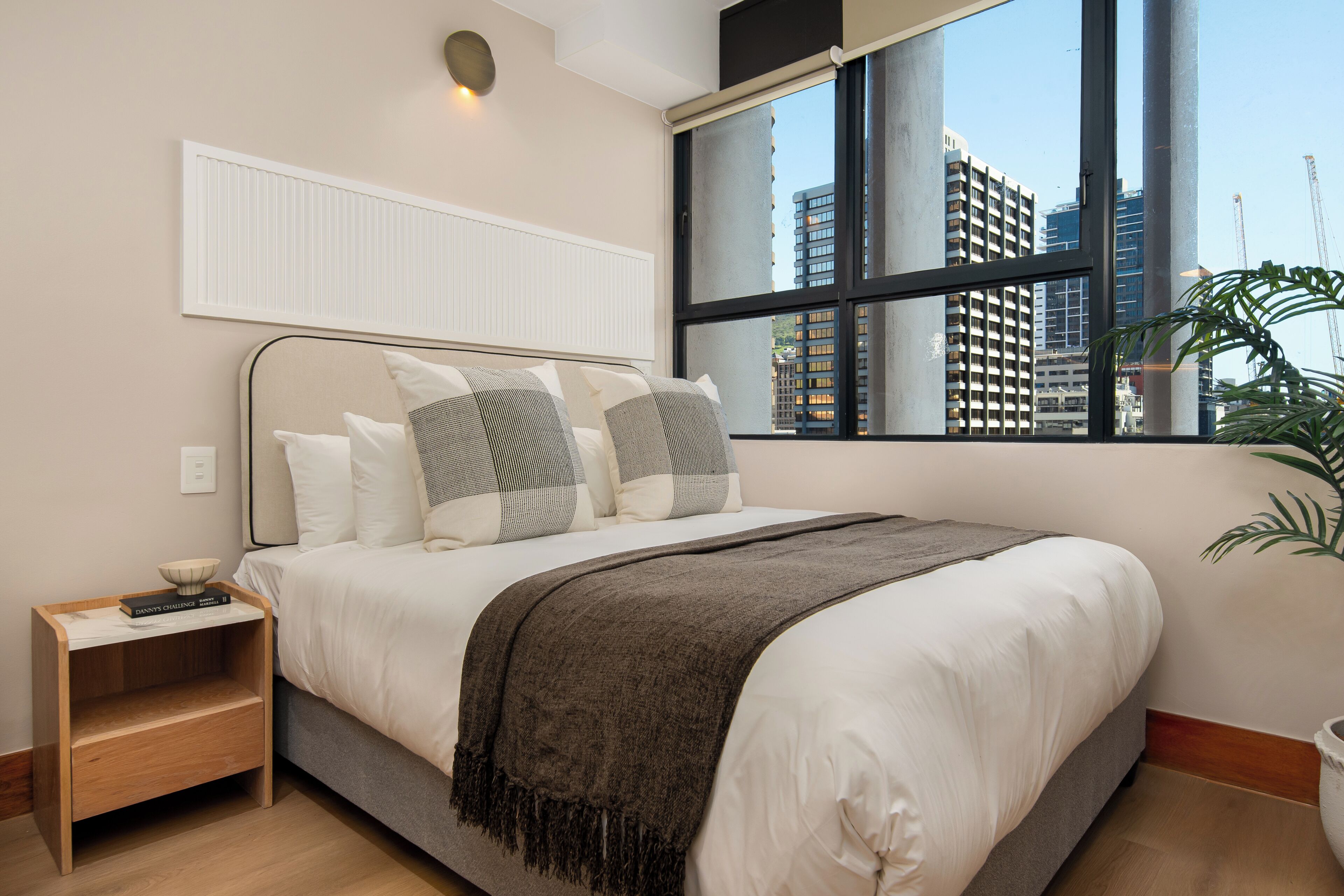 A modern bedroom with a large bed, a nightstand, a window with a view of buildings, and a potted plant.