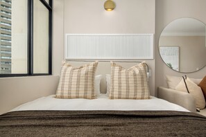 Habitación de hotel con cama, dos almohadas, una ventana, una lámpara de pared, un radiador y un espejo redondo.