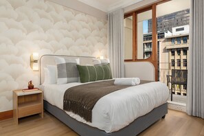 A hotel room with a large bed, a nightstand, a window with a view of buildings, and a patterned wall. - Charlie and the Chairman | Modern CBD Apartment (Cape Town)