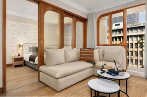 Ein modernes Hotelzimmer mit einer Couch, einem kleinen Tisch, einem Nachttisch und Blick auf die Stadt durch große Fenster.