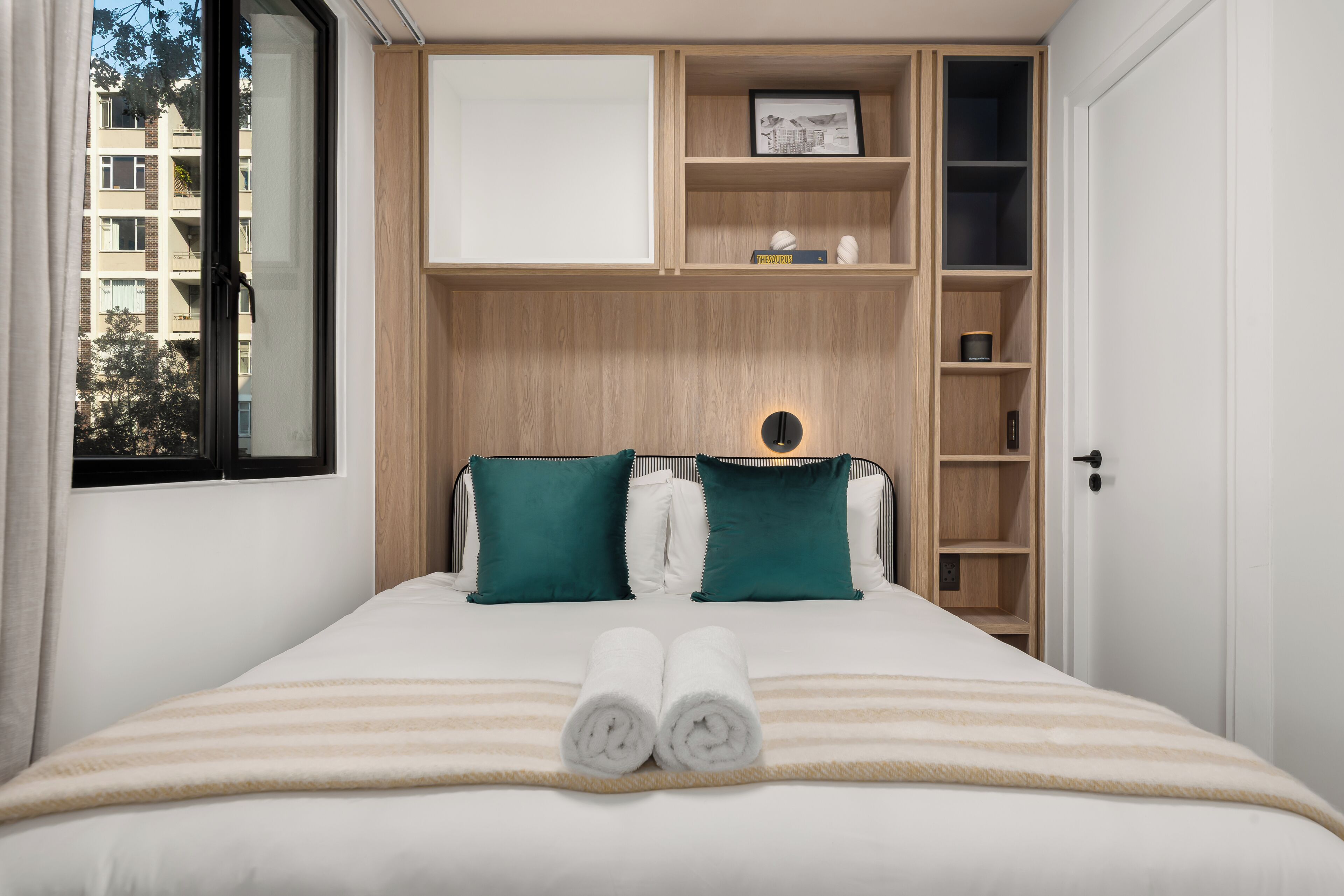 Un lit bien fait, avec deux oreillers, une table de chevet avec une lampe, une fenêtre et une bibliothèque intégrée avec un tableau encadré et un livre.