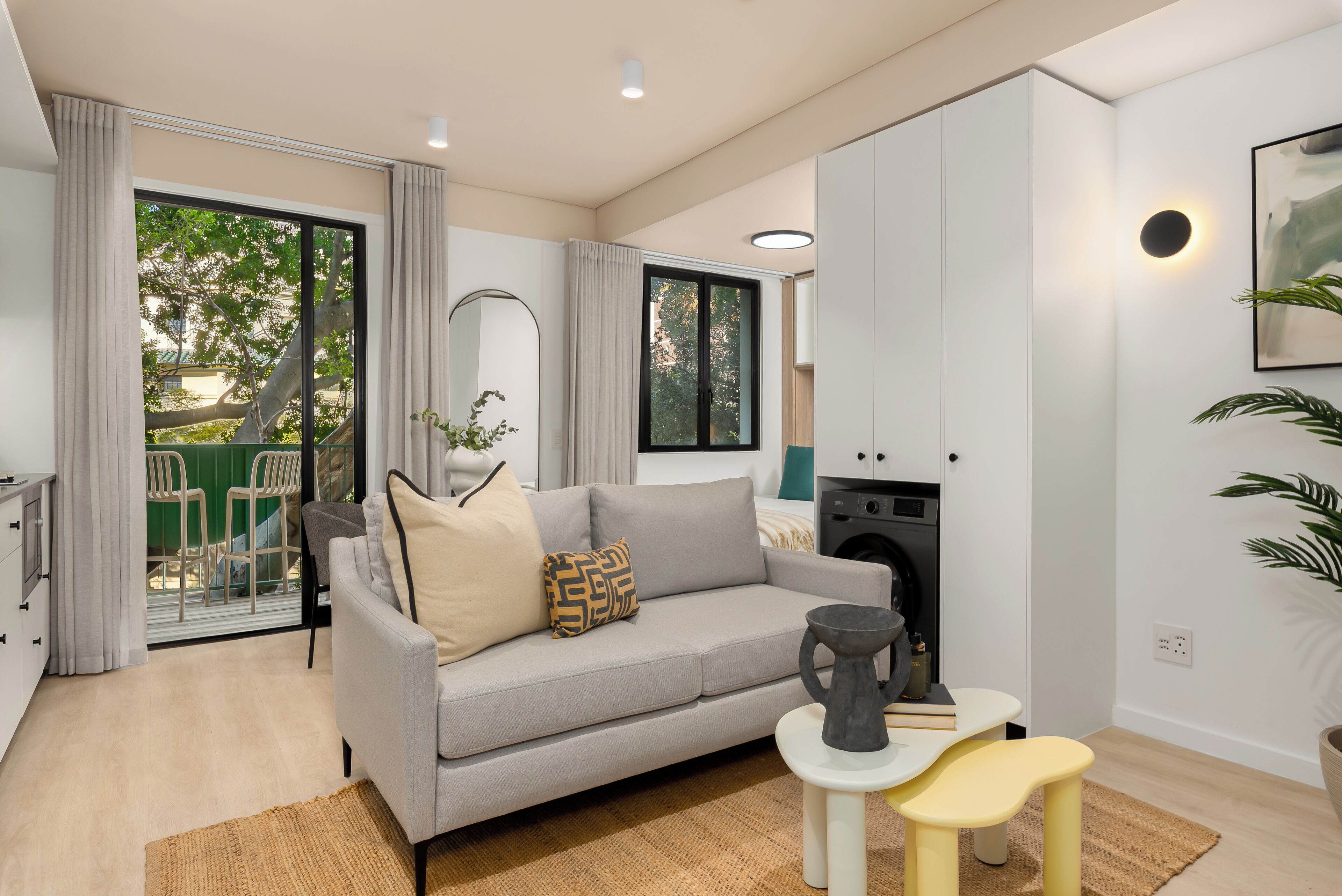 Una sala moderna con un sofá gris, una mesa de centro y una mesita auxiliar. Se ve una lavadora al fondo, y una zona verde al aire libre a través de la puerta de vidrio.