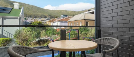 A balcony with wicker chairs and a small round table, overlooking a town with buildings and a hill in the distance.