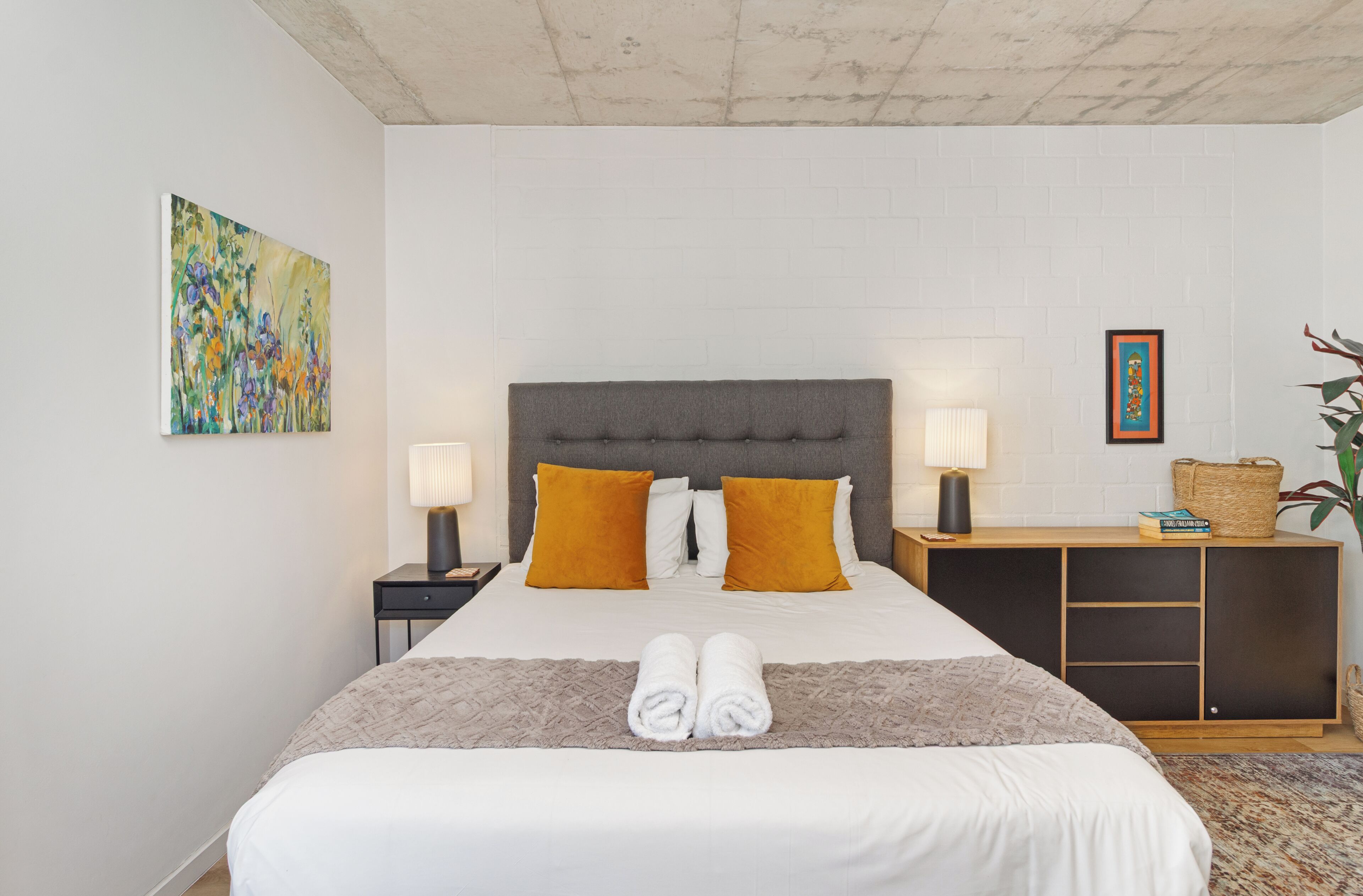 A hotel room with a large bed, two bedside lamps, a nightstand, a bedside table with a lamp, a small side table with a basket, and a framed picture on the wall.