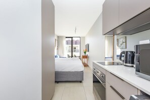 A modern hotel room with a bed, a kitchenette, and a view of the cityscape. - One Rosebank | Elevated Urban Comfort (Johannesburg)