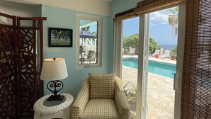 A room with a ceiling fan, a chair, a small table with a lamp, a picture on the wall, and a view of a swimming pool through large windows.