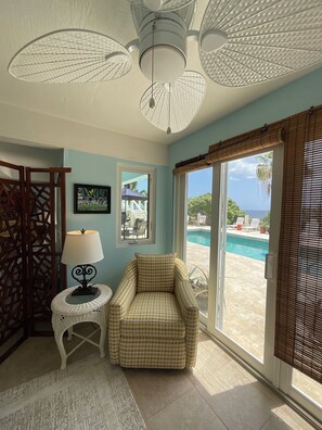 Une pièce dotée d’un ventilateur de plafond, d’une chaise, d’une petite table avec une lampe, d’un tableau au mur et offrant une vue sur une piscine grâce à de grandes fenêtres.