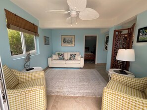 Ein Wohnzimmer mit einem weißen Sofa, gemusterten Sesseln, einem Deckenventilator, einem Fenster mit Jalousien und einer Tür zu einem anderen Raum.