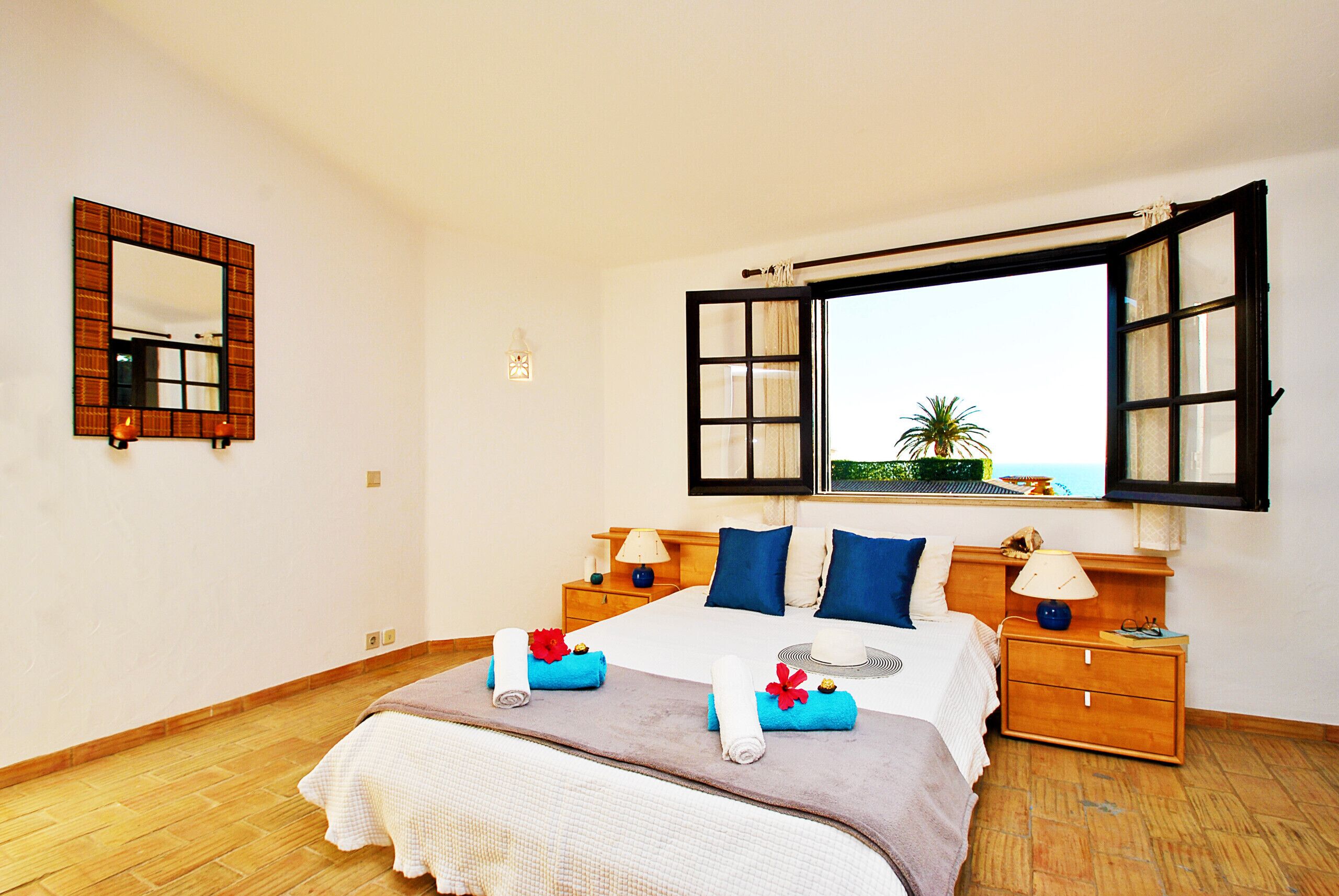 A bedroom with a bed, a nightstand, a lamp, a mirror, and an open window with a view.