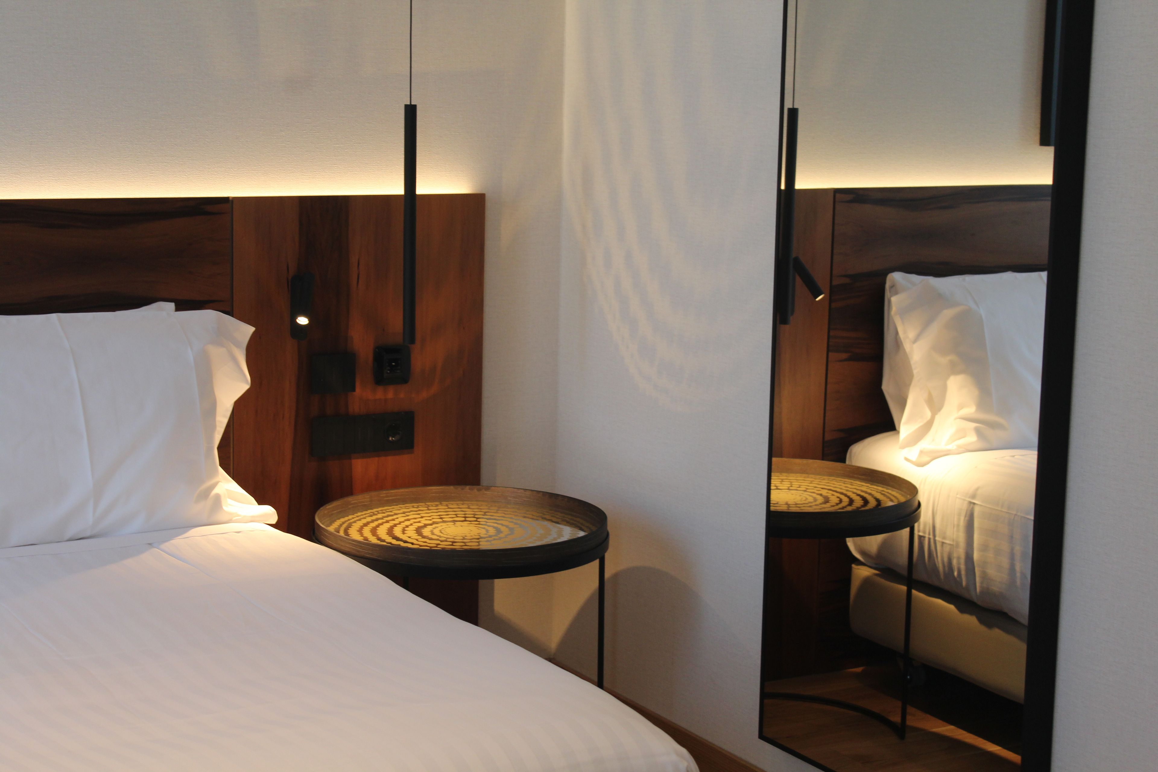Una habitación de hotel con cama, mesita de noche, espejo y lámpara de pared.
