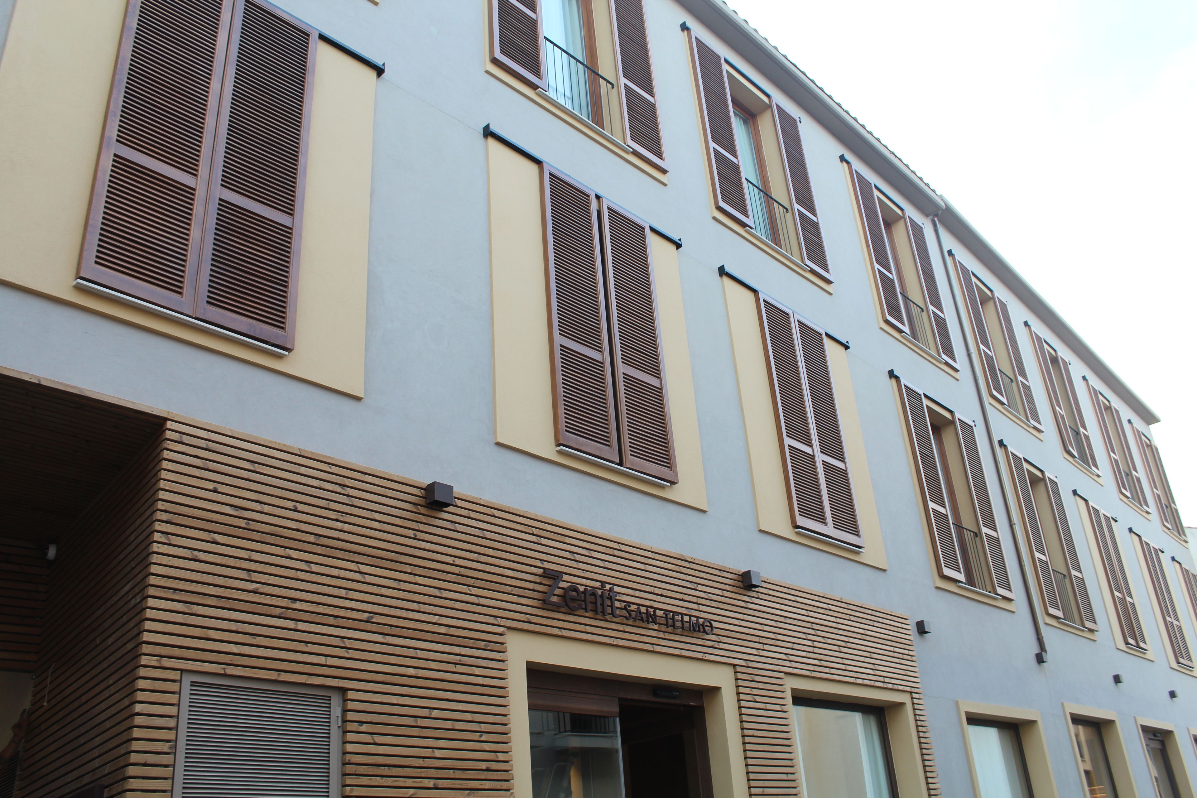Un edificio de varios pisos con fachada plana, que presenta una serie de ventanas con contraventanas y una entrada a nivel de calle con diseño de listones de madera.