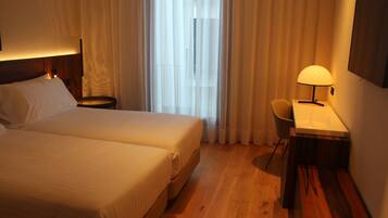 Habitación de hotel con dos camas, un escritorio, una silla, una lámpara y un amplio ventanal con cortinas.