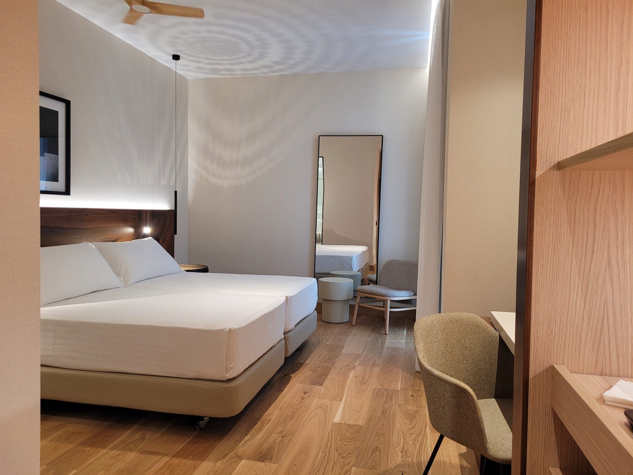 Una habitación de hotel con cama, una silla, un escritorio, un espejo y un ventilador de techo.
