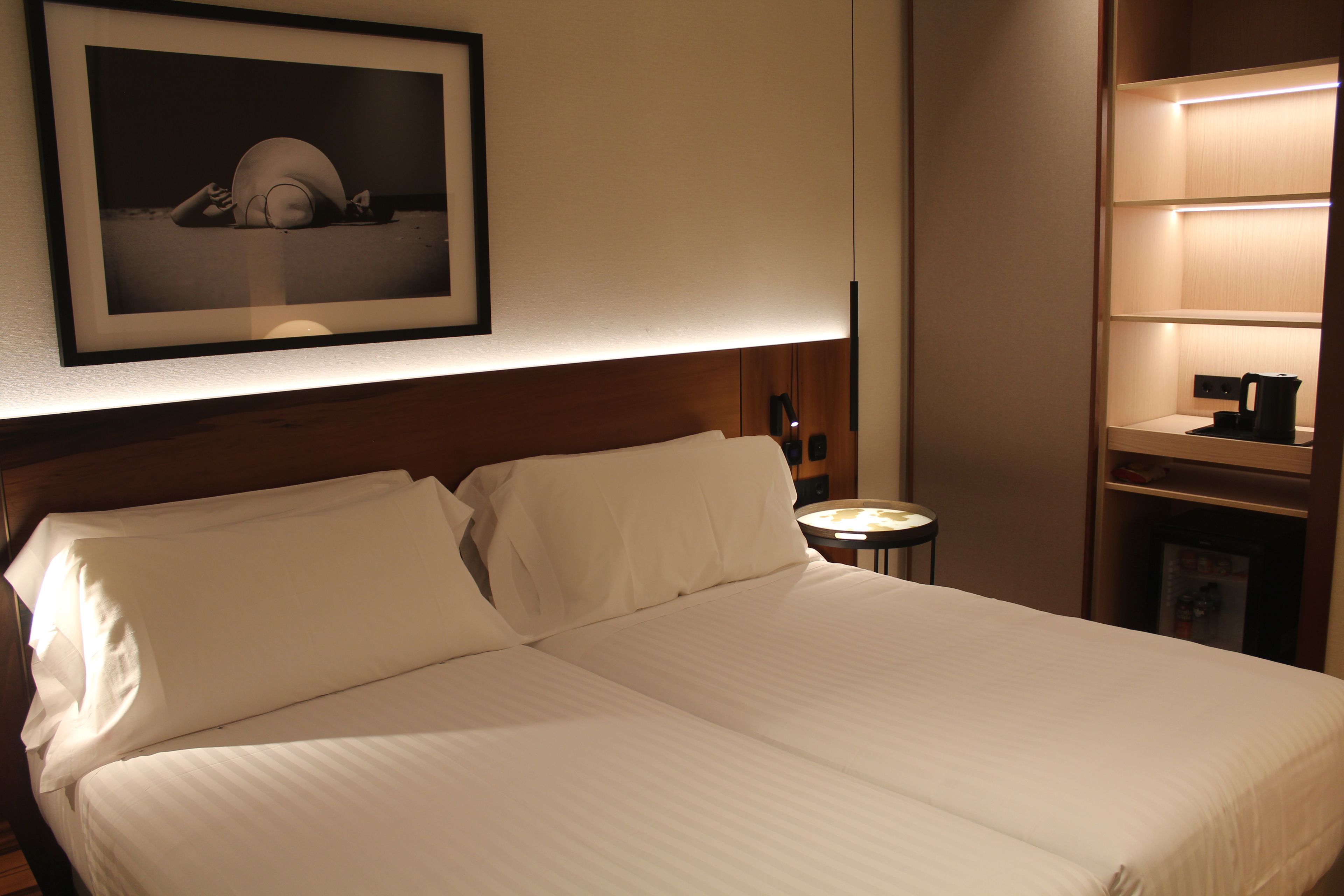 Una cama bien hecha en una habitación de hotel, con un cuadro enmarcado en la pared, una mesita de noche con lámpara y un estante con hervidor.