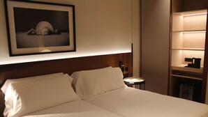 Een net opgemaakt bed in een hotelkamer met een ingelijst beeld aan de muur, een nachtkastje met een lamp en een plank met een waterkoker.