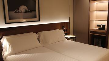 Una cama bien hecha en una habitación de hotel, con un cuadro enmarcado en la pared, una mesita de noche con lámpara y un estante con hervidor.