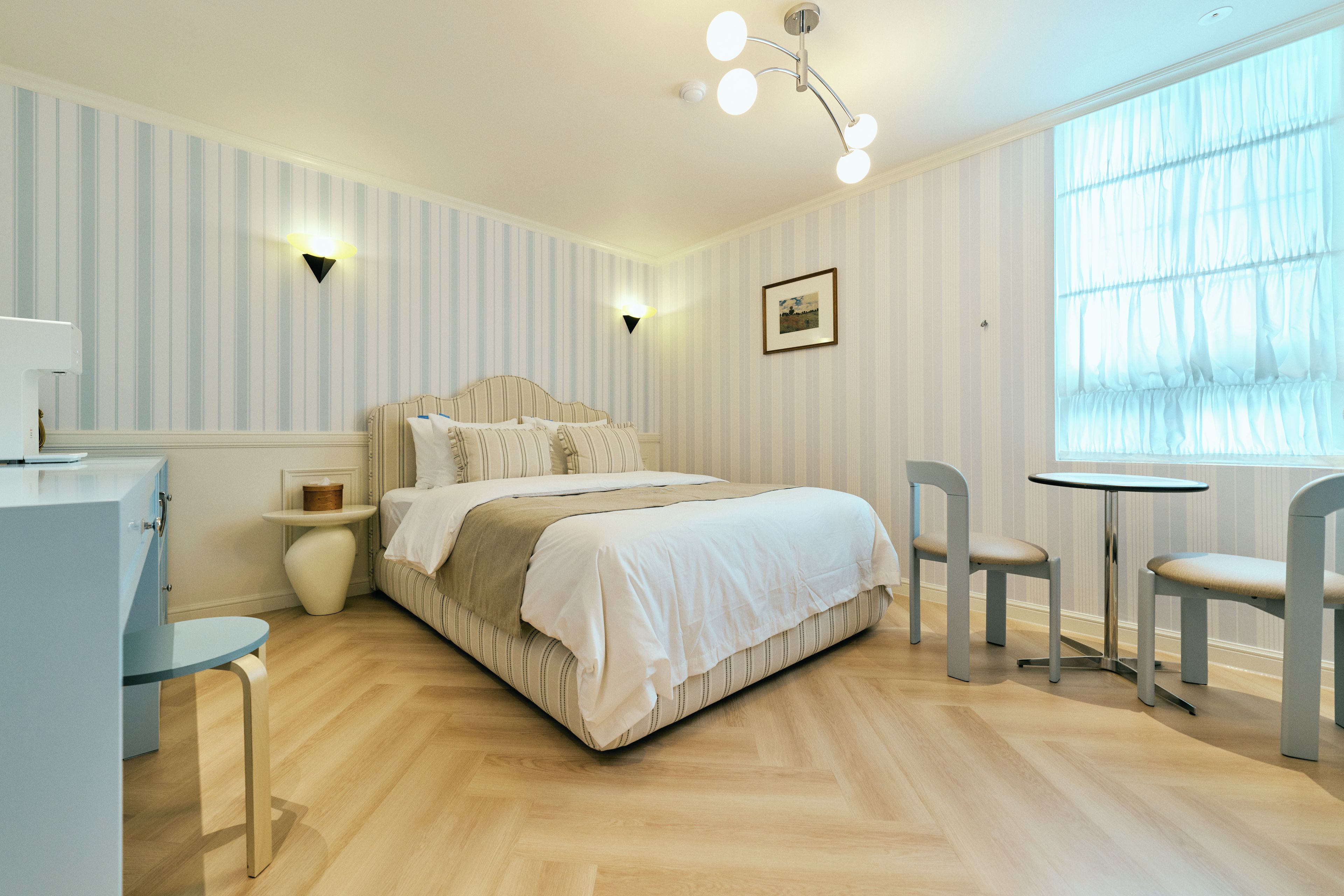 Un dormitorio con cama, mesitas de noche, escritorio, silla, una pequeña mesa y ventana con cortinas.