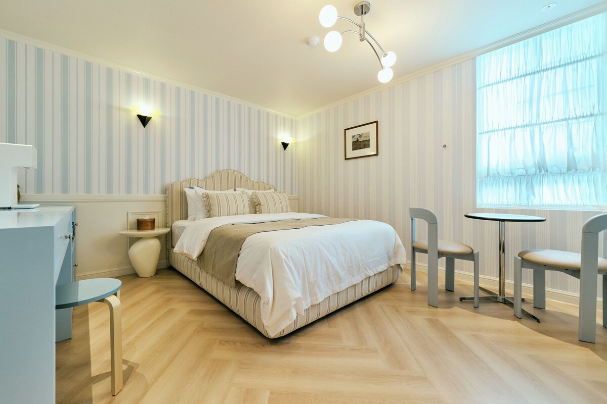 Un dormitorio con cama, mesitas de noche, escritorio, silla, una pequeña mesa y ventana con cortinas.