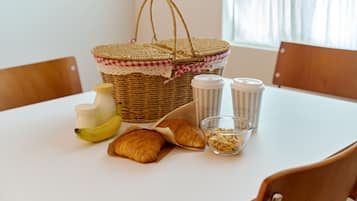 Una mesa con una cesta de mimbre, dos botellas de condimentos, un plátano, dos croissants y un pequeño recipiente de vidrio con cereal.