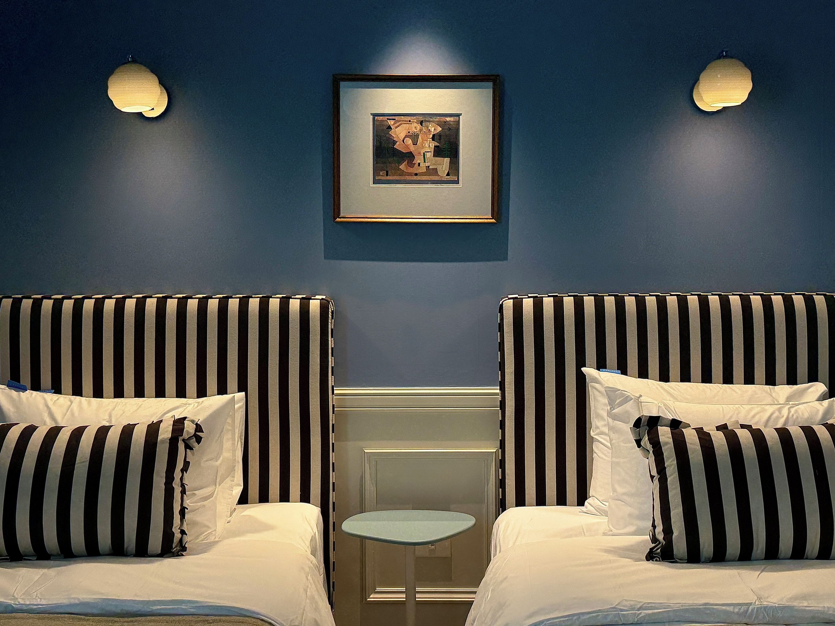 A hotel room with two beds, each with striped bedding, a small round table, and a framed picture on the wall.