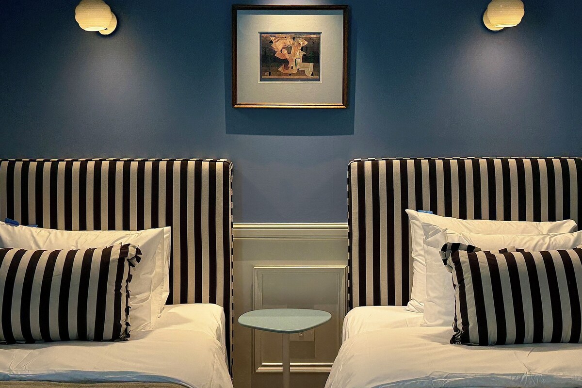 A hotel room with two beds, each with striped bedding, a small round table, and a framed picture on the wall.