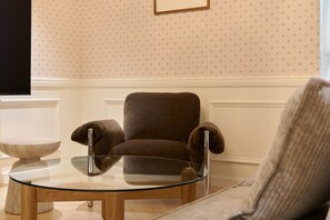 Una habitación con un sillón marrón, una mesa de centro de vidrio y una mesita auxiliar blanca.