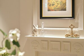 Una repisa blanca con una obra de arte enmarcada, un conejo de cerámica blanca, una botella de vidrio transparente y dos jarras de vidrio ornamentadas.