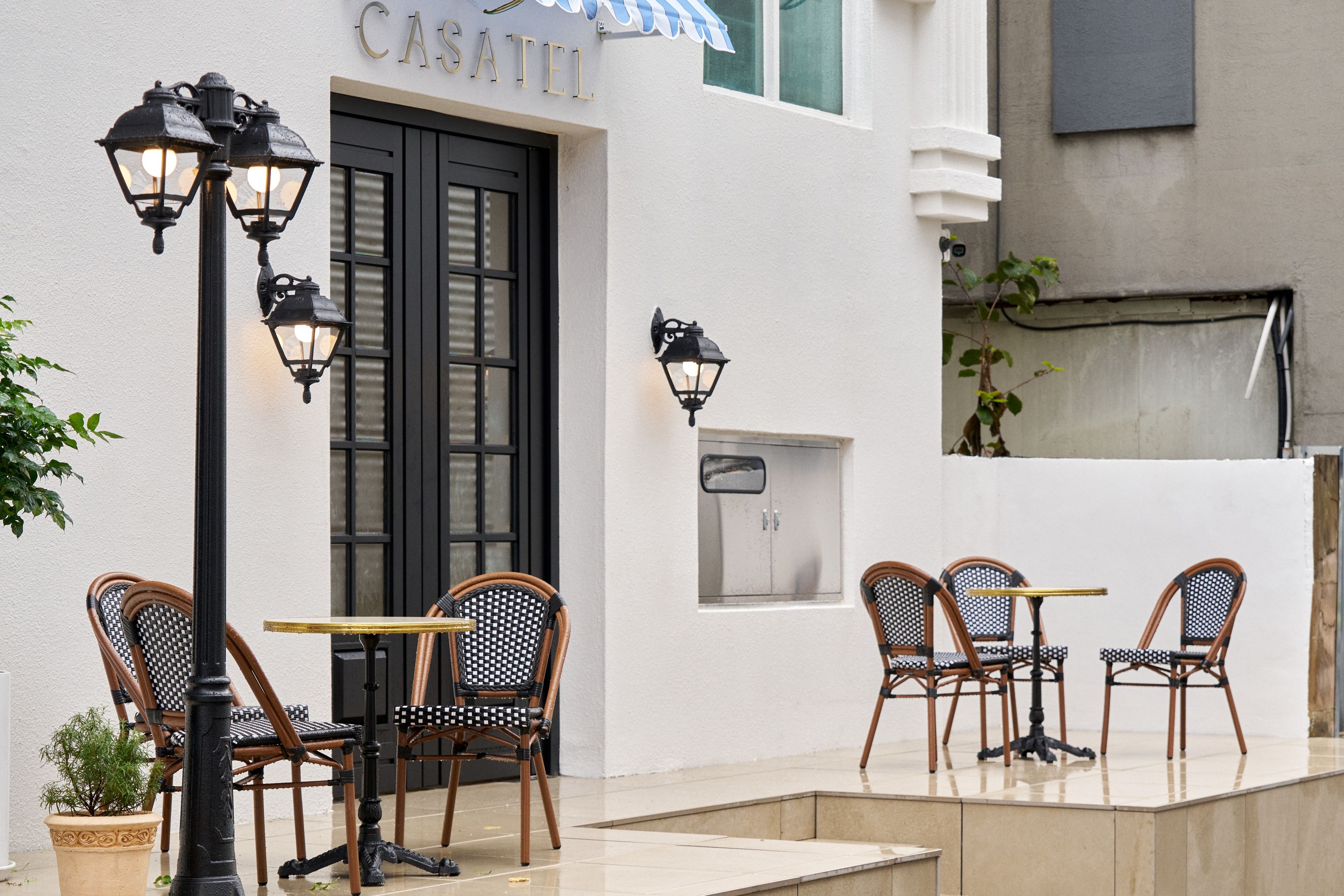 Un área de estar al aire libre con sillas y mesas de mimbre, una puerta negra con el nombre 'CASATEL' en la parte superior y faroles fijados a la pared.