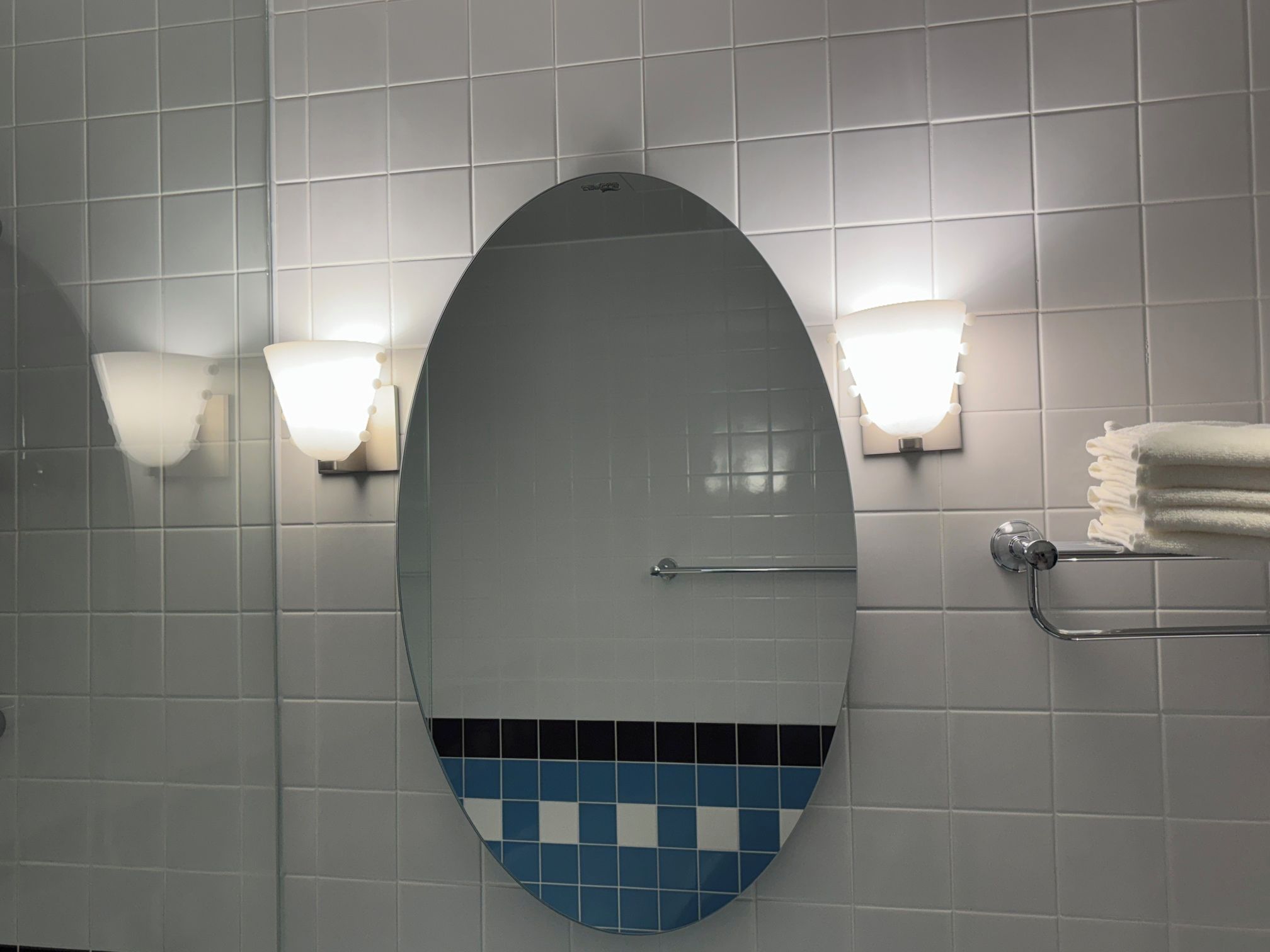 Een badkamer met een grote ovale spiegel, twee wandlampen, een handdoekhouder en een accentmuur met blauw en wit tegelwerk.