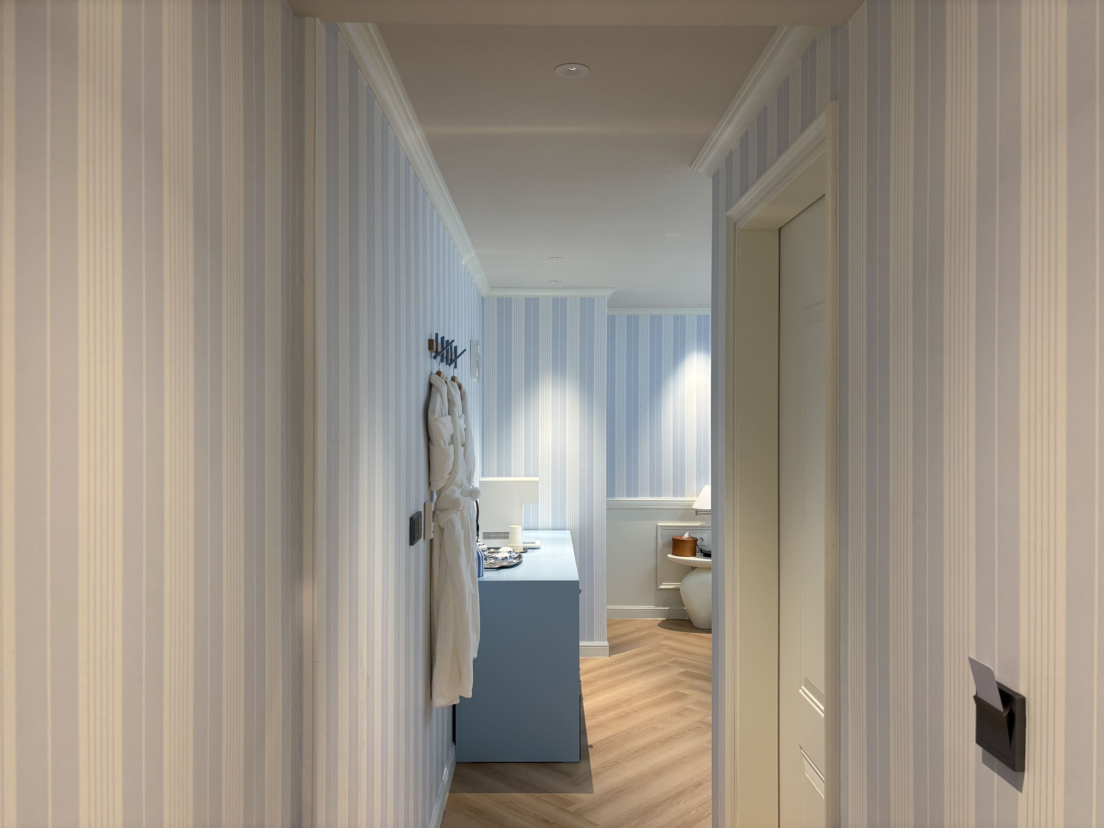 A hallway with striped wallpaper, a blue room with a bed and bedside table, and a door on the right.