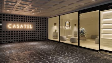 Una entrada de hotel moderna con piso de baldosas de piedra, fachada de vidrio y el nombre 'CASATEL' iluminado en la pared.