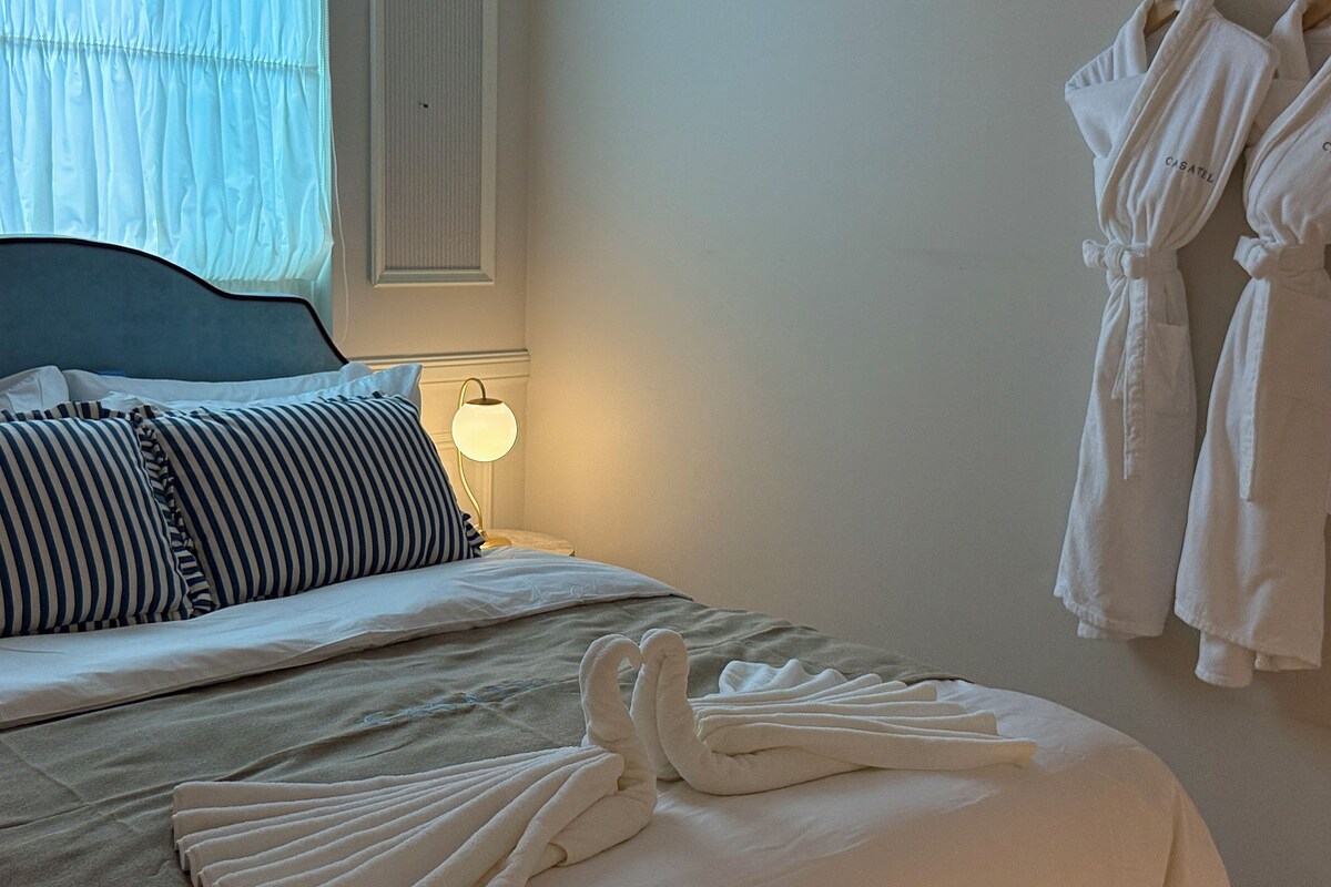 Una cama bien tendida con almohadas a rayas, una lámpara de noche y un juego de batas colgadas en la pared.